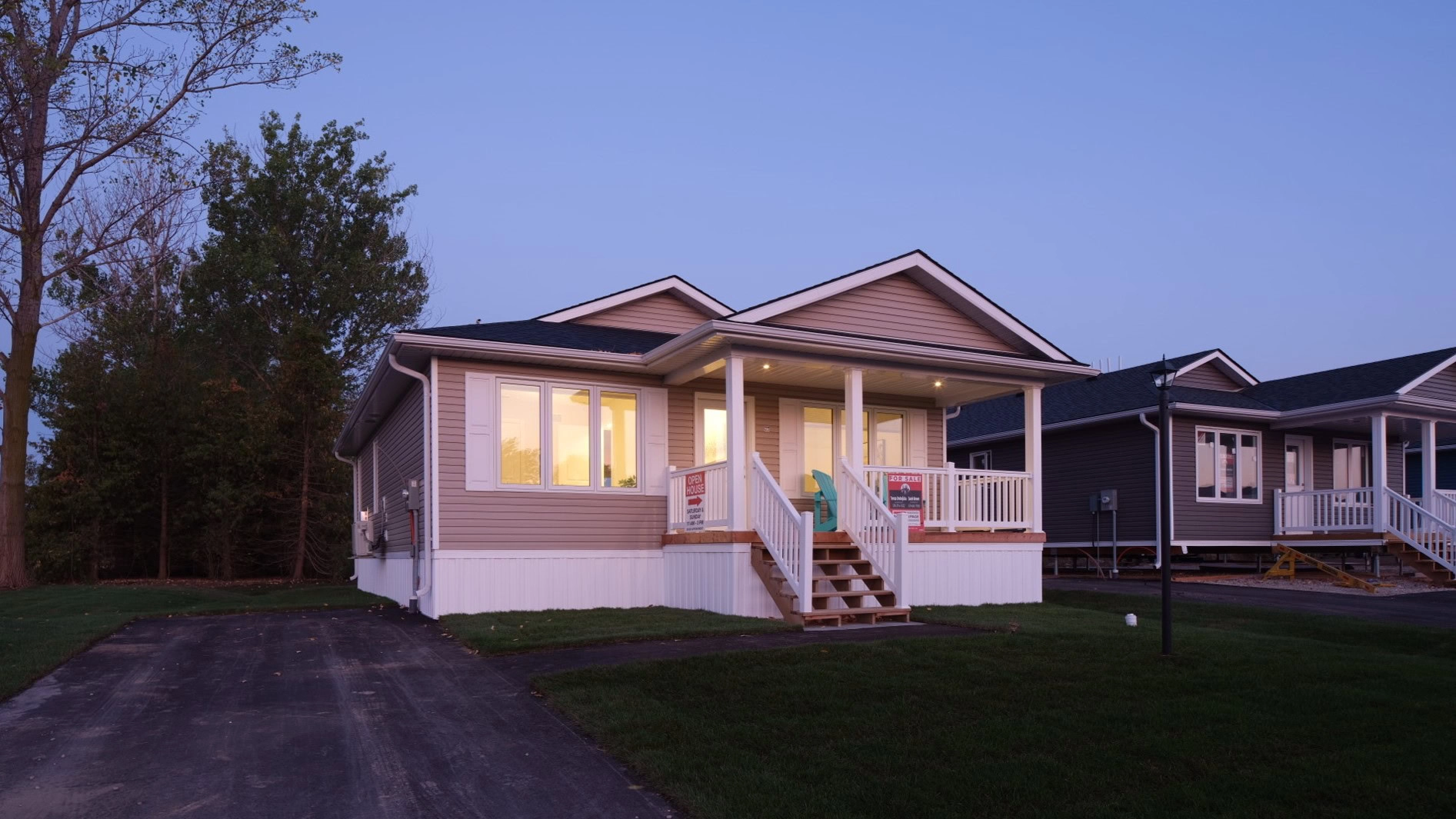 A single-story house at dusk with lights on inside, surrounded by a grassy yard and trees.