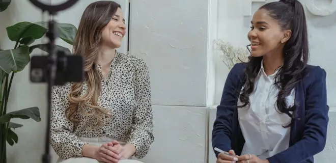 Two people are sitting and smiling at each other in a bright, indoor setting. A camera is set up in front of them, suggesting an interview or recording session.