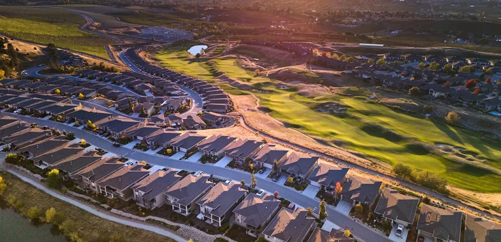 An aerial view of a suburban neighborhood with a row of houses alongside a landscaped golf course. The scene is bathed in warm, evening sunlight.