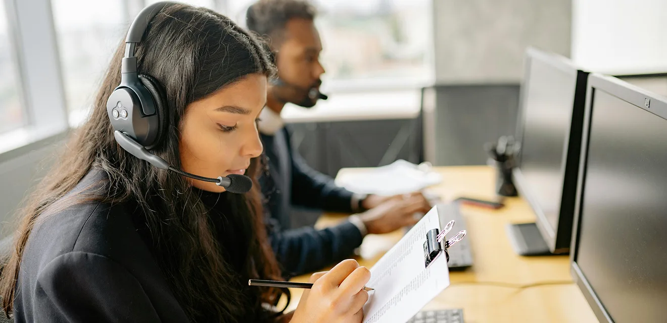 Two people working at a desk with computers, wearing headsets.