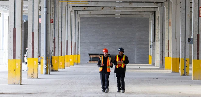 Two people wearing safety vests and helmets are walking through an expansive, empty industrial warehouse.