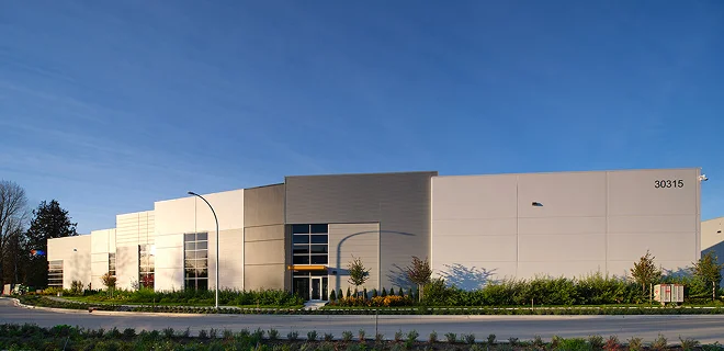A large, modern industrial building with a flat roof and minimalistic design. It is surrounded by greenery and set against a clear blue sky.