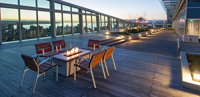A rooftop patio with chairs around a fire pit, offering a view of the city skyline and a river at sunset. The area is modern and well-lit.