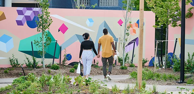 A couple walks holding hands through a park with a colourful mural.