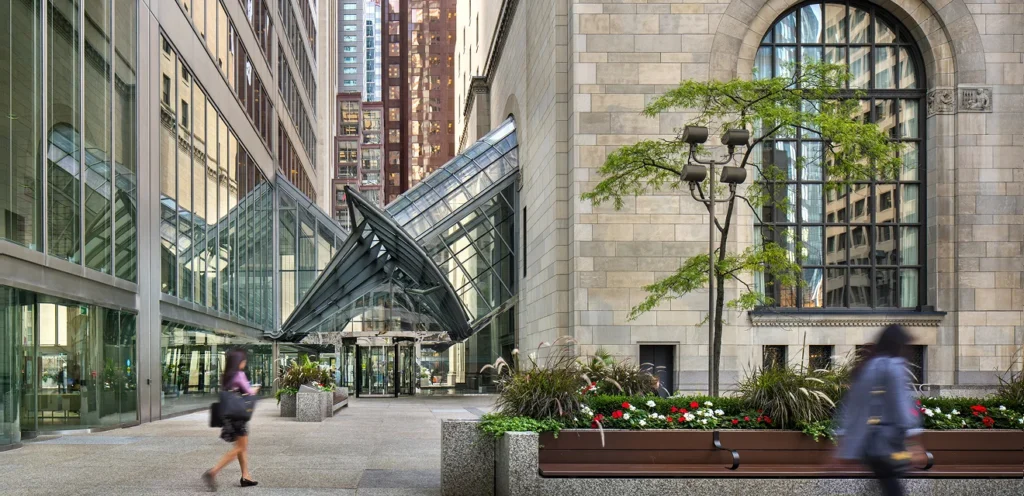 A city scene featuring modern glass structures adjacent to a historic building. People are walking along the sidewalk, and the area is enhanced with greenery and planters.