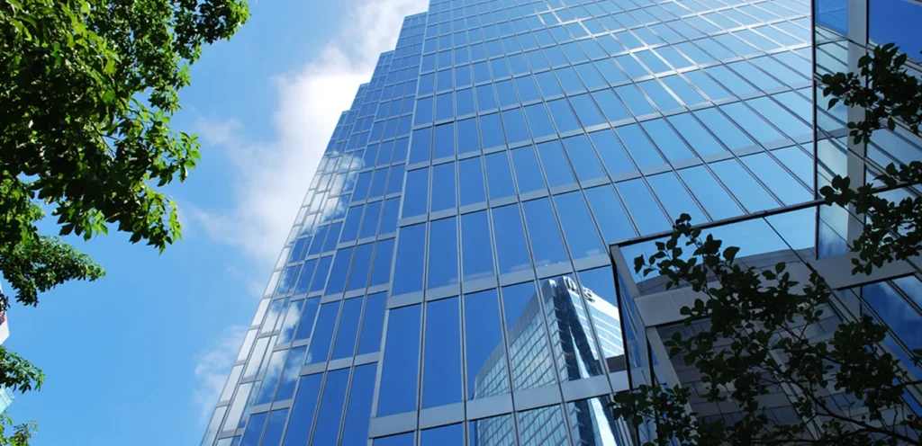 An upward view of a modern skyscraper with a reflective glass facade, set against a backdrop of a blue sky with a few clouds. Trees frame the scene.