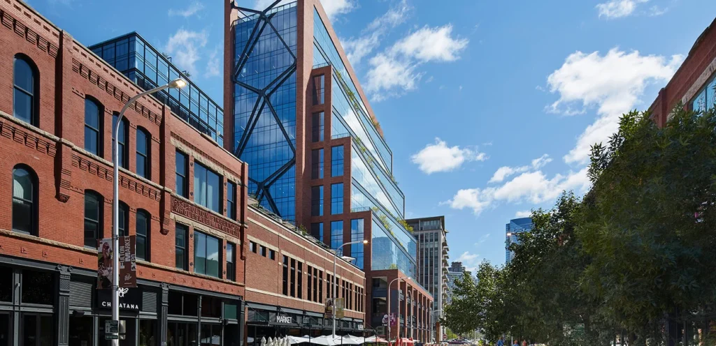 A street view featuring a mix of historic brick buildings and modern glass architecture, under a bright blue sky with scattered clouds. Trees line the sidewalk.