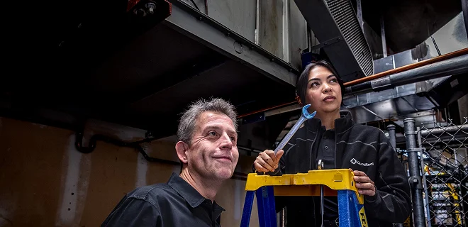 Two people looking off camera. One of them is on a ladder holding a wrench.