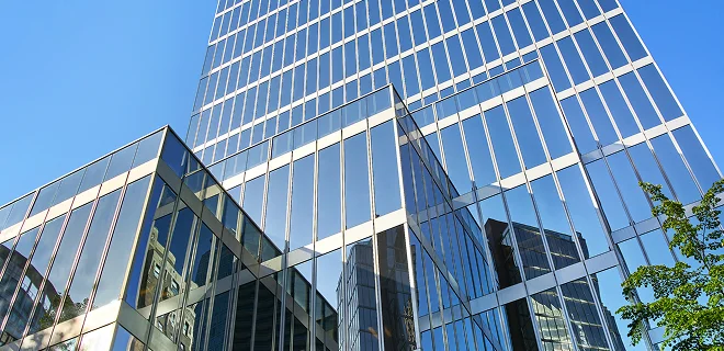 Aerial view looking down at a vehicle driving on a narrow road through a forest. A tall glass office buidling with some trees in the foreground.