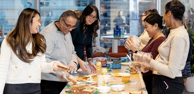 A group of people gathered around a table, laughing and interacting, possibly during a casual cooking or baking session. The atmosphere is lively and joyful.