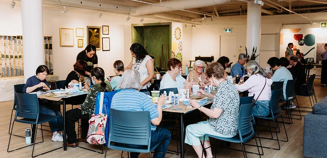 A group of people sitting at tables in a brightly lit room, engaged in various activities or discussions. The setting is open and welcoming, suggesting a communal gathering or workshop.