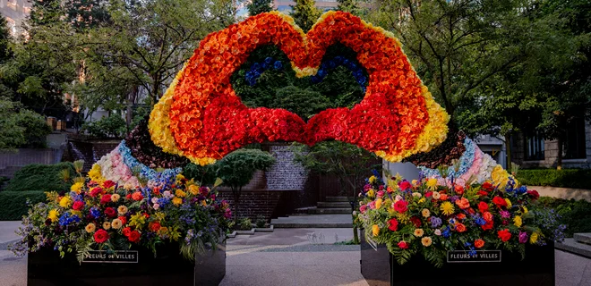 A vibrant flower installation shaped like a heart, made up of red, orange, and yellow blooms, set in a garden area surrounded by greenery.
