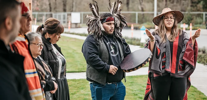 A group of people gathered outdoors, some wearing traditional attire and holding instruments. It appears to be a cultural or ceremonial event.