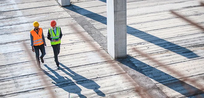 Two construction workers wearing safety vests and helmets are walking across a large, open construction site. They are in conversation, with shadows cast on the ground.