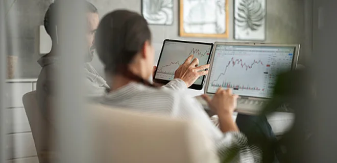 Two people are collaborating in an office, looking at a computer screen displaying charts and graphs. One person is pointing at the screen. The setting is professional and focused.