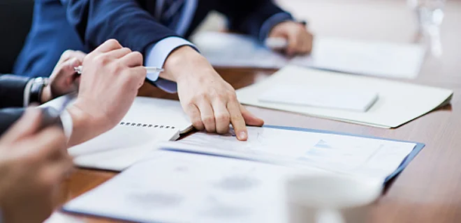 Close-up of business professionals reviewing documents at a meeting. Hands are pointing at charts and graphs on papers spread across a table.