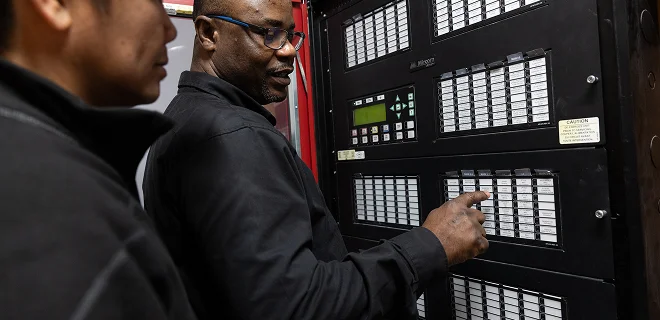 Two technicians are examining and discussing a control panel with switches and indicators in a technical setting.