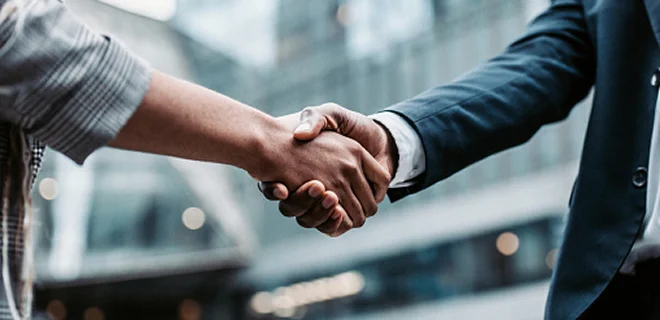A close-up of a handshake between two individuals, set in an urban environment. One person is wearing a suit.