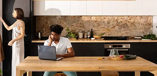 Person using a laptop at a kitchen table, while a person stands near a cabinet