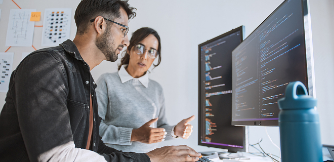 L’image montre deux personnes travaillant ensemble à un bureau avec deux écrans d’ordinateur.