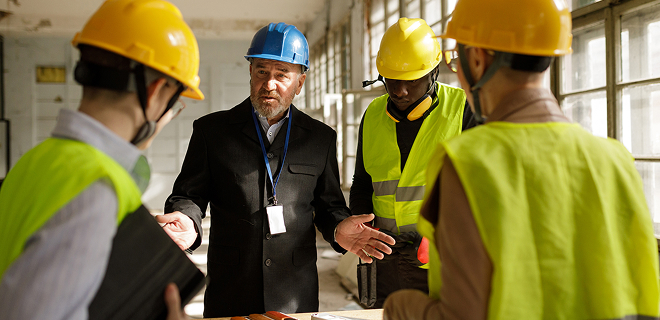 L’image montre un groupe de personnes sur un chantier de construction portant des casques de sécurité et des gilets de haute visibilité.
