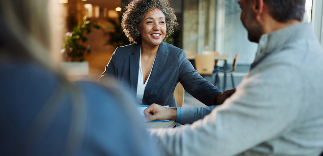 L’image montre une personne qui sourit et discute avec deux autres personnes, qui sont assises à une table.
