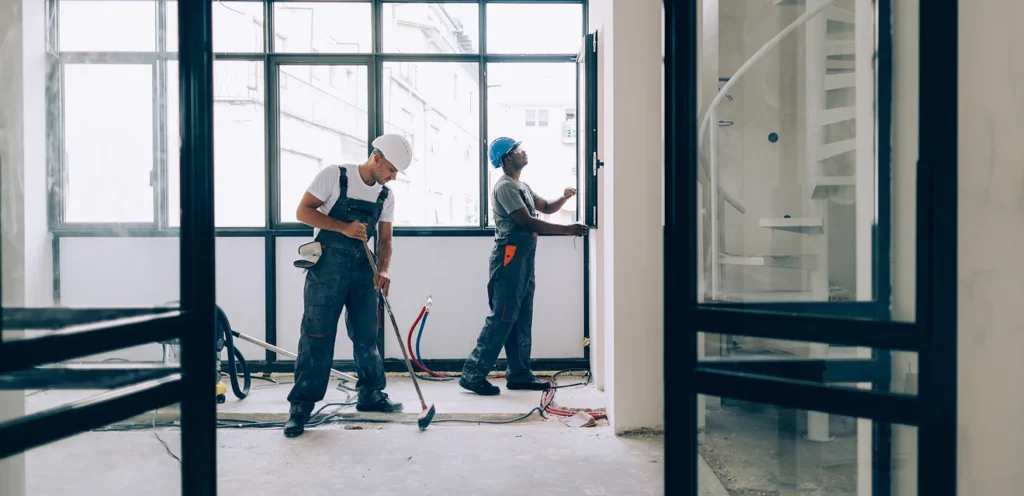 Deux travailleurs de la construction portant un uniforme et un casque de sécurité travaillent à l’intérieur d’un immeuble avec de grandes fenêtres.