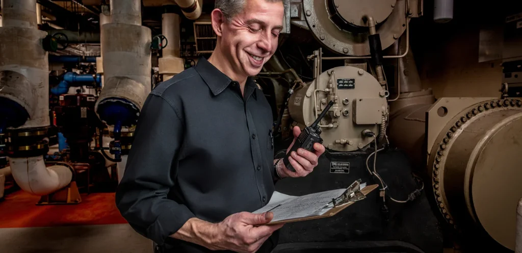 A technician smiling while working in an industrial setting, holding a clipboard and a radio. The background features machinery and pipes.