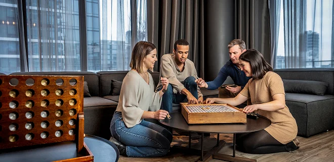 A group of four people sitting in a cozy, modern living space with large windows, playing a board game together. There's a relaxed and friendly atmosphere.