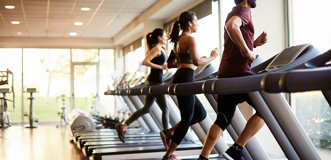 People are running on treadmills in a well-lit gym with large windows, creating an energetic and focused atmosphere.