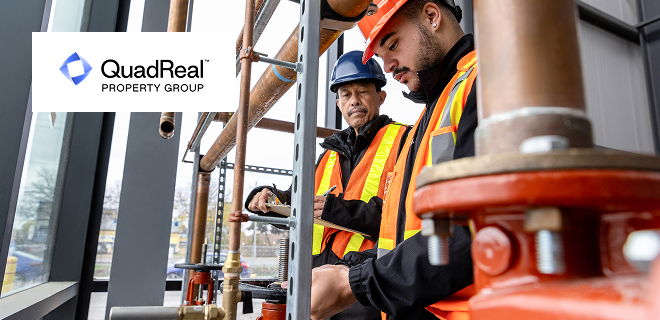 Two individuals wearing safety gear and helmets are inspecting or working on industrial piping inside a building.