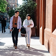Two people walking along a sunny city street lined with buildings and trees, engaged in conversation.