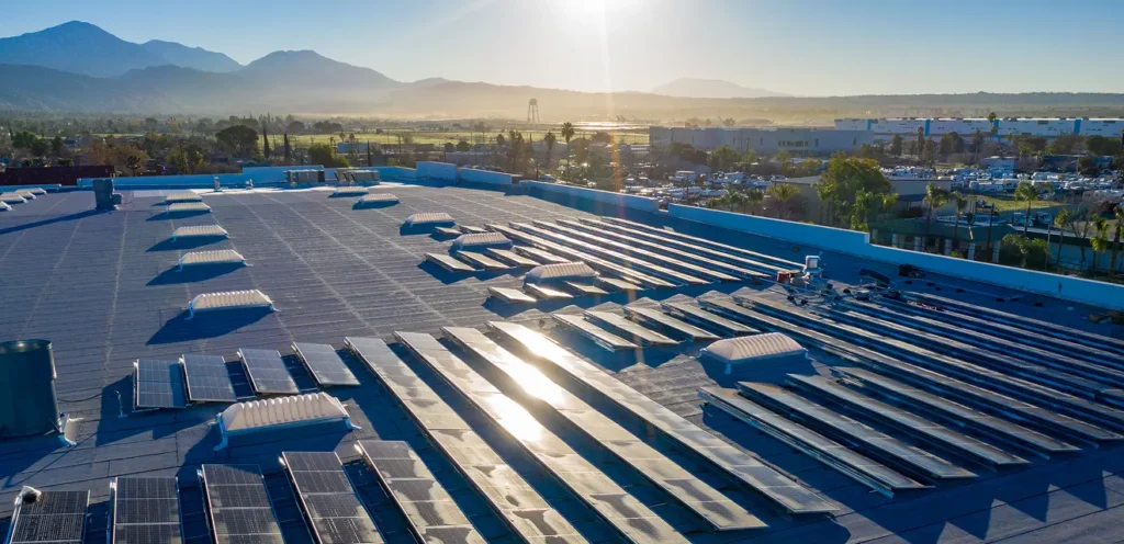 A rooftop covered with solar panels, overlooking a scenic landscape with mountains in the background and the sun low in the sky.