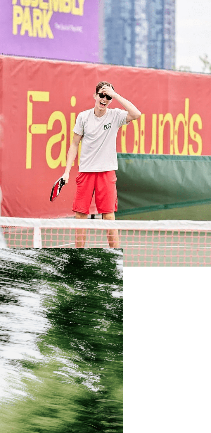 Une personne debout sur un terrain de tennis, tenant une raquette et se touchant la tête, portant des lunettes de soleil et une tenue décontractée. L’arrière-plan comporte une bannière colorée.