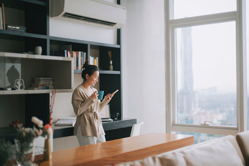 A woman holding a mug and looking at a tablet stands behind a table near a window.