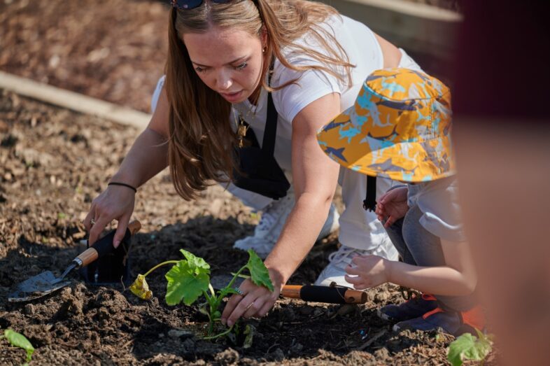 Une personne et un enfant font du jardinage ensemble, plantant une jeune plante dans le sol. L’enfant porte un chapeau coloré.