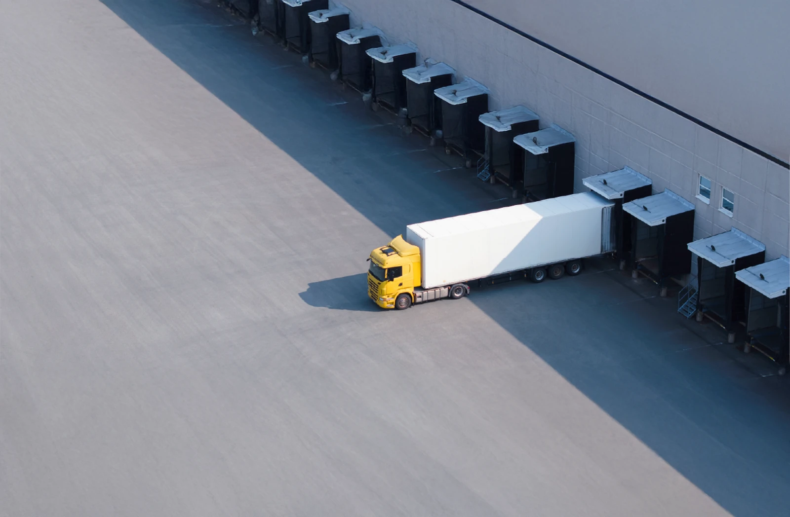 Aerial view of a yellow semi-truck with a white trailer parked at a loading dock of an industrial warehouse