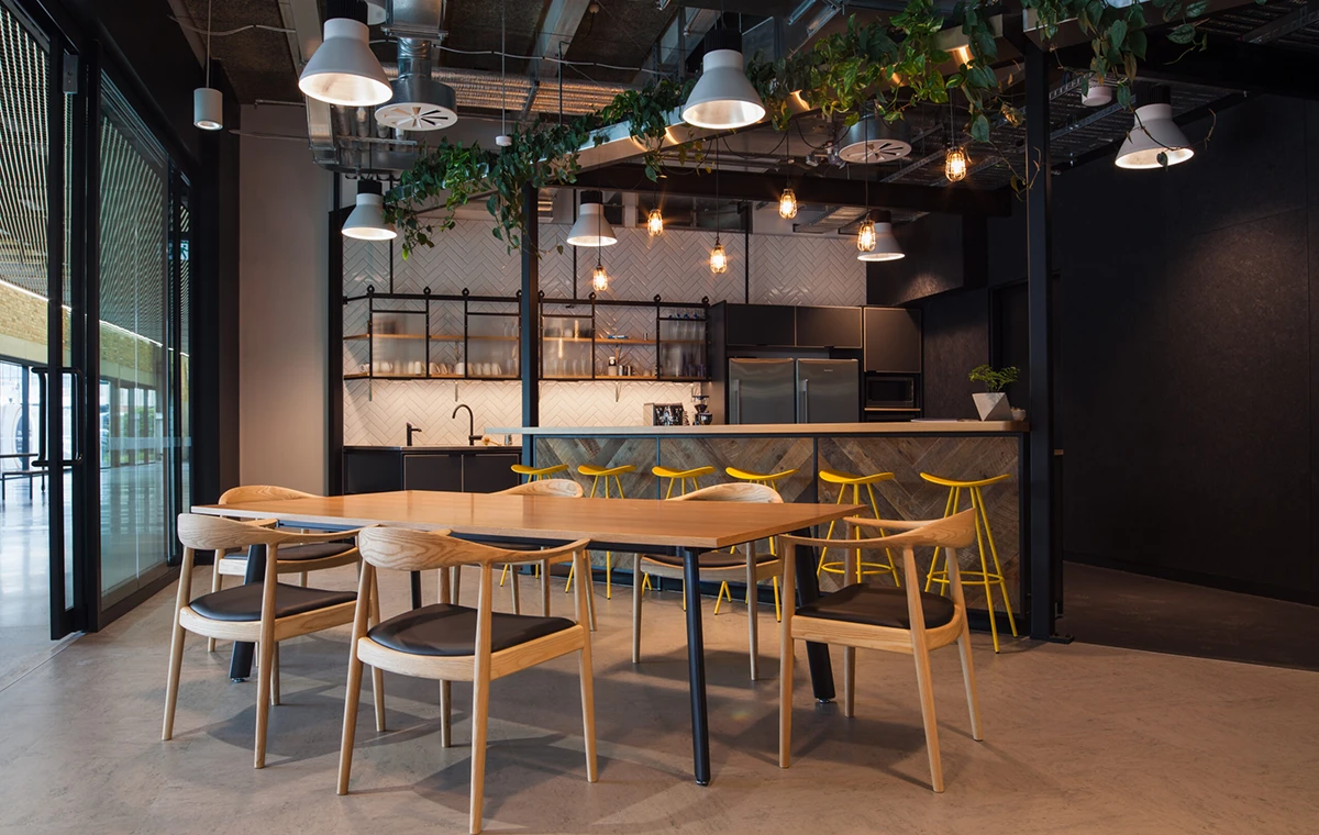 A modern kitchen and dining area with a wooden table and six chairs in the foreground. The kitchen features a long counter with four yellow bar stools, hanging pendant lights, and open shelves with glassware. There are plants hanging from the ceiling, adding a touch of greenery to the space.