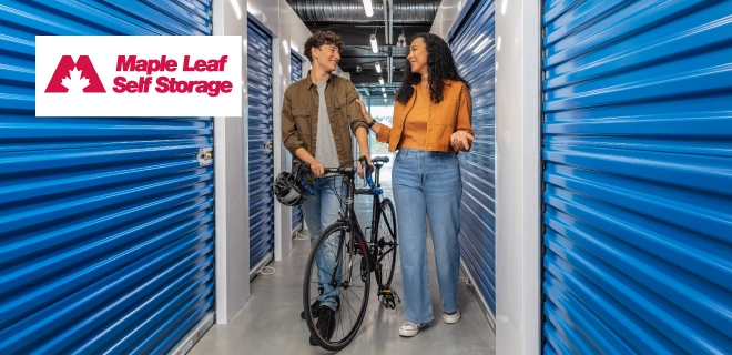 Two casually dressed people stand in a bright, modern self-storage hallway with blue roll-up doors. One holds a bike and helmet; the other gestures mid-conversation. A red maple leaf logo for Maple Leaf Self Storage appears in the top left.