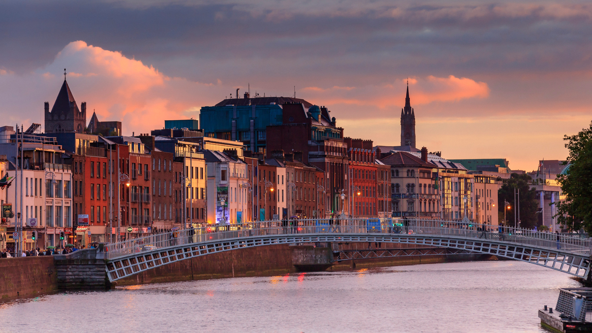 A scenic city view at sunset, featuring a pedestrian bridge over a river, colorful buildings along the waterfront, and historic spires in the background.