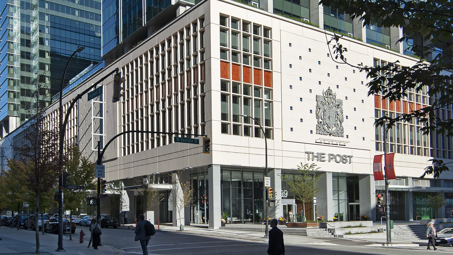 Street view of The Post building in downtown Vancouver, featuring a modern facade with vertical window panels, the building name ‘The Post’ and a large crest on the exterior wall. Pedestrians are crossing the intersection in front, and surrounding high-rise glass towers are visible in the background.