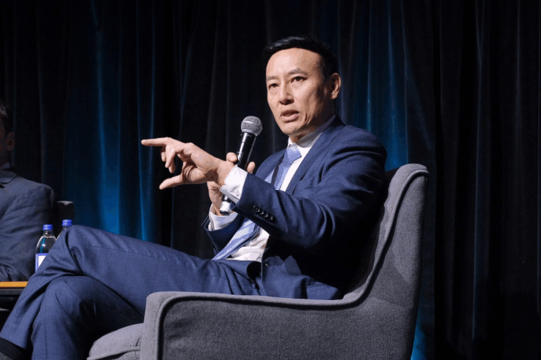 Person in a blue suit seated on a gray armchair, holding a microphone and gesturing with one hand during a panel discussion.
