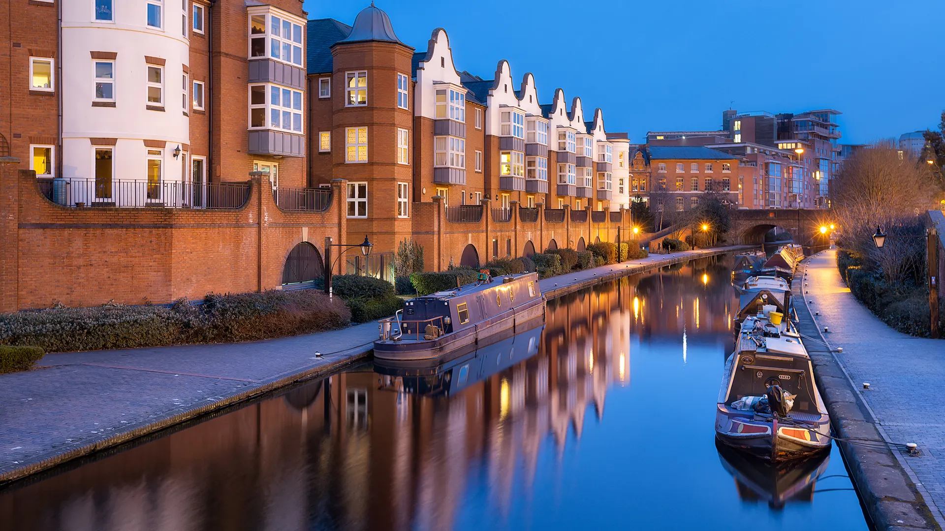 Immeubles résidentiels en brique le long d’un canal au crépuscule, avec des péniches amarrées se reflétant dans l’eau.