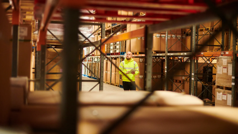 Travailleur en veste haute visibilité inspectant l’inventaire dans une allée d’entrepôt remplie de boîtes empilées et d’étagères.
