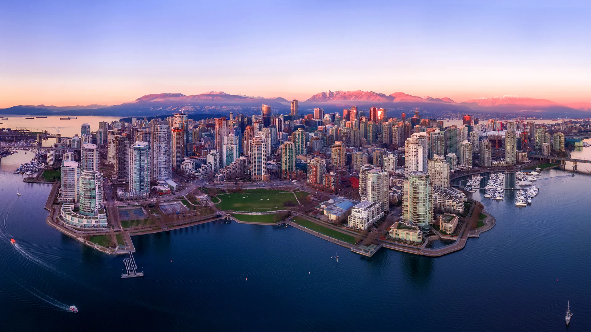 Vue aérienne du centre‑ville de Vancouver au lever du soleil, montrant des gratte‑ciel entourés d’eau, de marinas et de chaînes de montagnes en arrière‑plan.