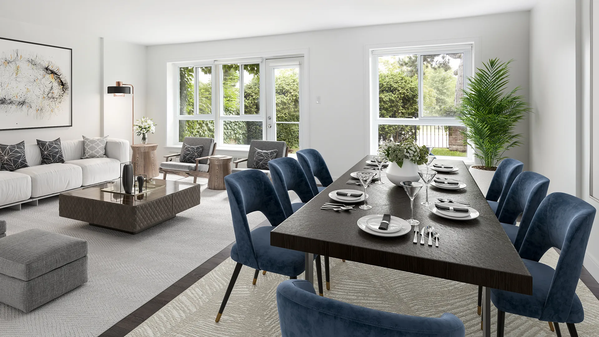 A bright open‑concept living and dining room with a long dining table set for a meal, blue chairs, a sofa seating area, and large windows overlooking greenery.