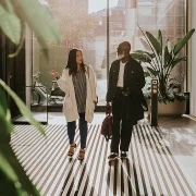 Deux personnes marchent ensemble dans une rue ensoleillée, entourées de bâtiments et d’arbres.