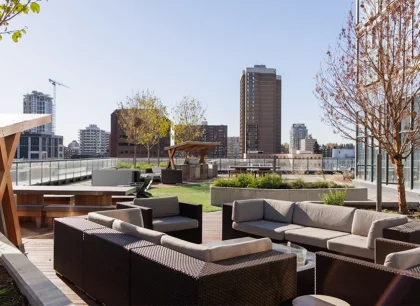 A rooftop terrace with comfortable seating, overlooking a cityscape with tall buildings. The area includes greenery and is bathed in sunlight.