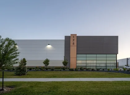 A modern industrial building with a flat roof and large windows. It is surrounded by a well-maintained lawn and small trees, under a clear evening sky.
