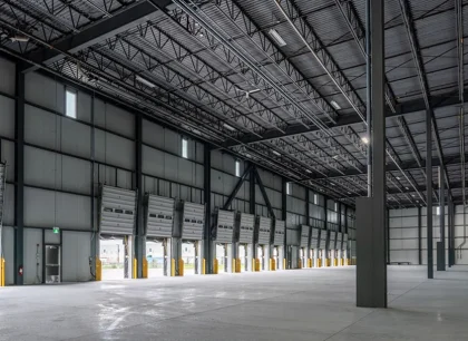 A spacious, empty industrial warehouse with high ceilings, large metal beams, and concrete flooring, illuminated by natural light from windows.